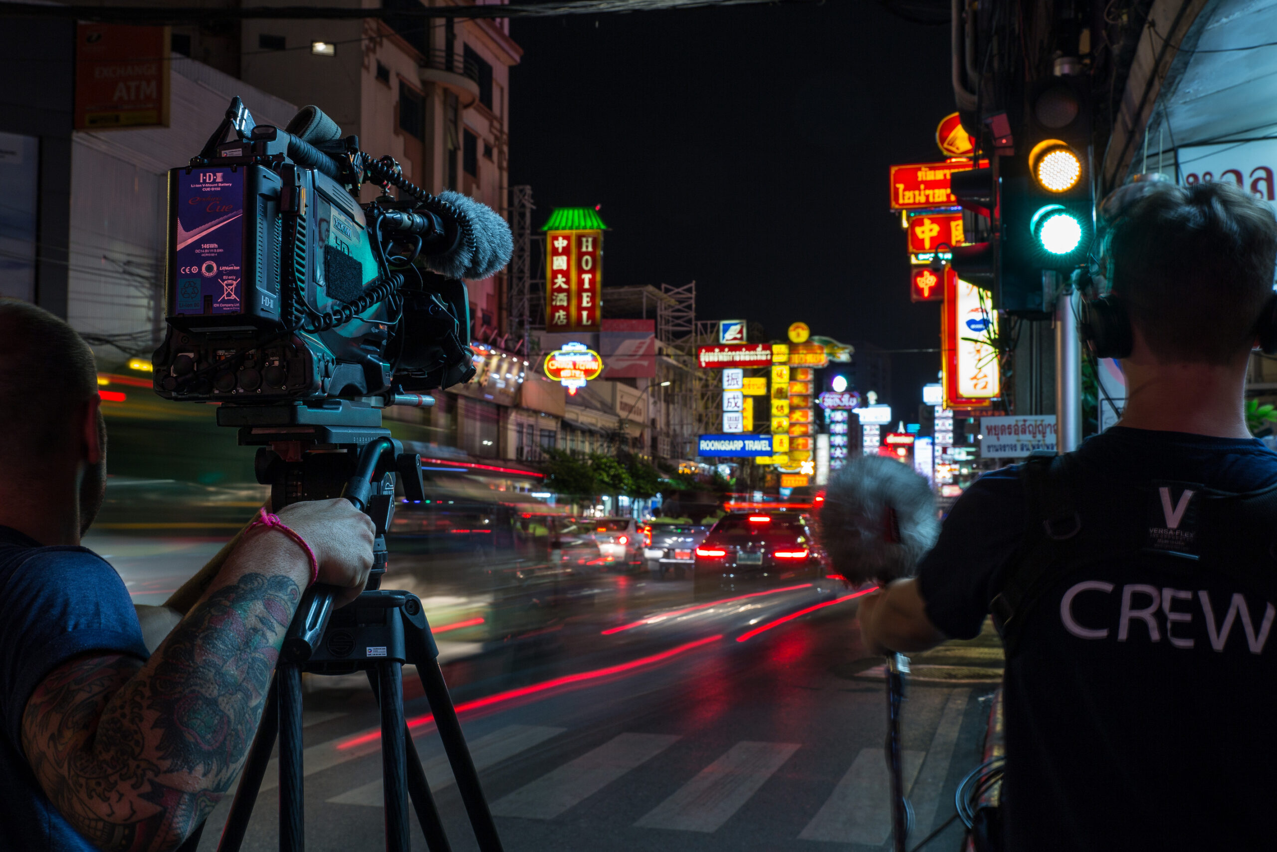 A professional camera crew for hire in Thailand films a dynamic long-exposure shot of a busy street at night in Bangkok, capturing the energy of the city with neon signs and light trails from traffic.
